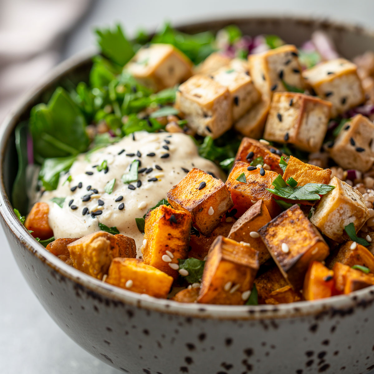 A ceramic bowl with a grain salad topped with cubed sweet potato and tofu with a tahini sauce.